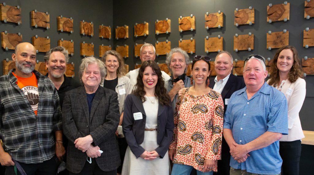 A group of eleven people standing together in a gallery with wooden block art mounted on the dark wall behind them.