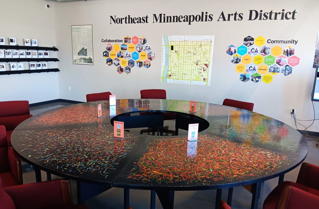 A large circular conference table with a colorful splattered paint design surrounded by chairs in a room with wall displays labeled Collaboration and Community for the Northeast Minneapolis Arts District.