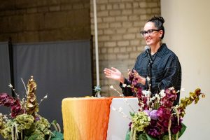 A woman with glasses speaking at a podium decorated with flowers and an orange draped cloth.