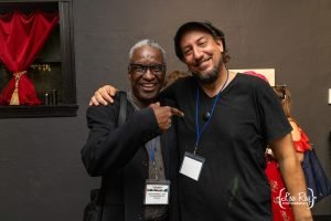 Two men smiling and posing for a photo, with the man on the left pointing at the man on the right; both wear conference lanyards.