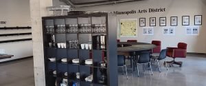 A black shelving unit filled with glassware stands in front of a round meeting table, with a Minneapolis Arts District map and framed prints displayed on the back wall.