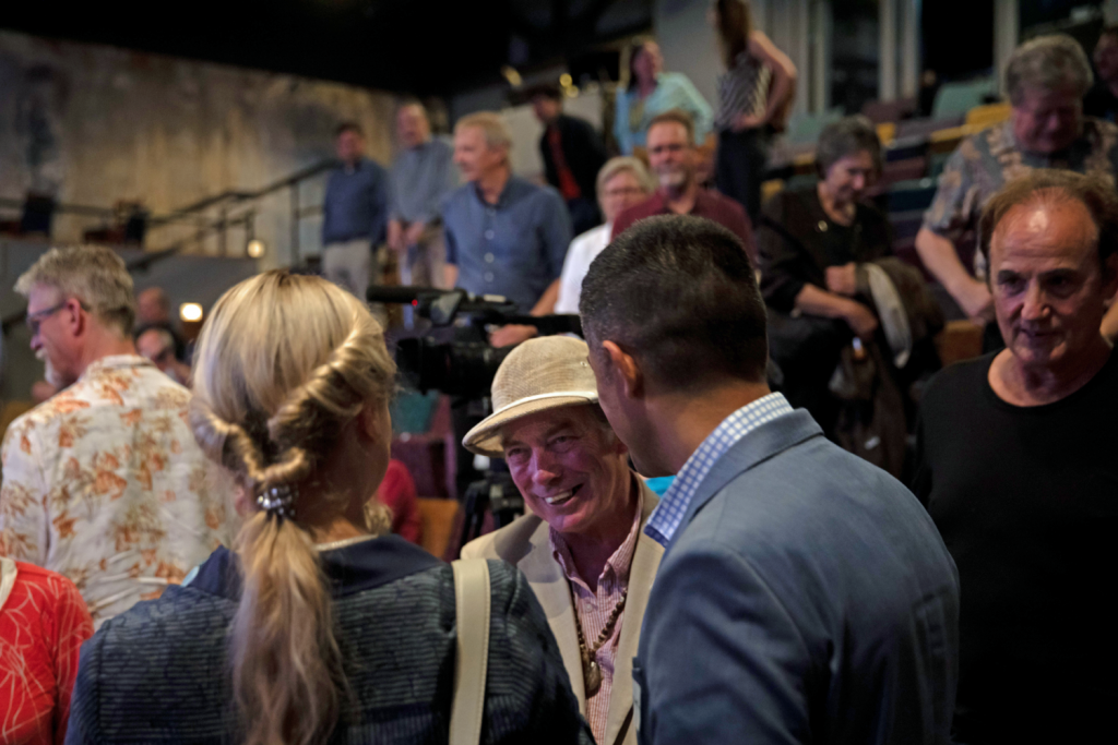 A group of people socialize in a theater or auditorium setting, with a smiling man in a beige hat at the center of a conversation.