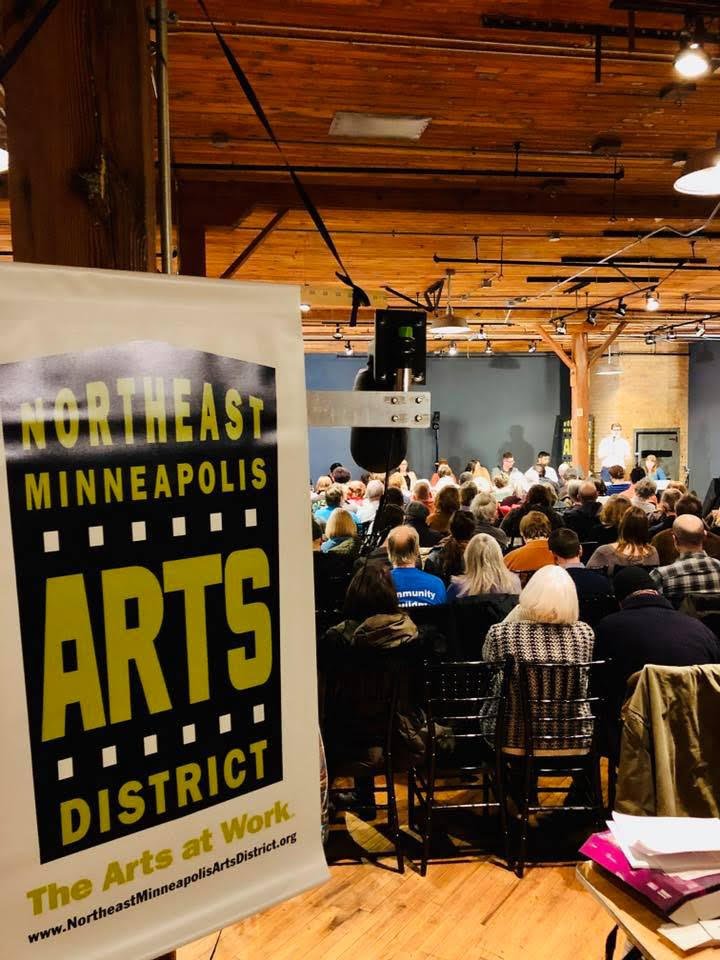 A sign for the Northeast Minneapolis Arts District stands before an audience seated in a wood-beamed room watching a presentation.
