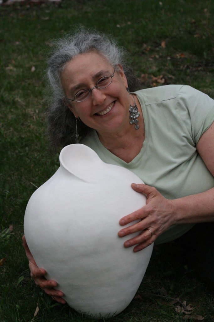 A smiling woman with gray hair holding a large, round, white ceramic vessel.