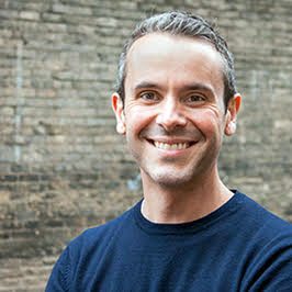 A smiling man in a dark blue shirt standing in front of a brick wall.