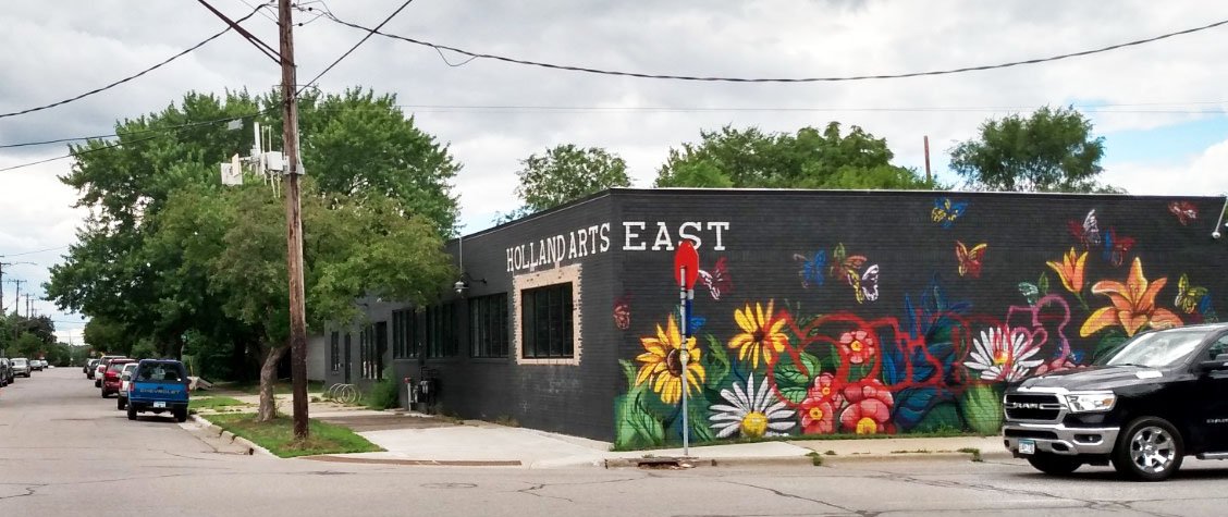 A black building labeled HOLLAND ARTS EAST featuring a colorful floral and butterfly mural on its exterior corner.