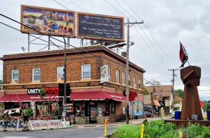 A brick building housing a Unity Foods market with murals and protest signs, featuring a billboard with religious quotes above and a large raised fist sculpture topped with a Pan-African flag in the foreground.