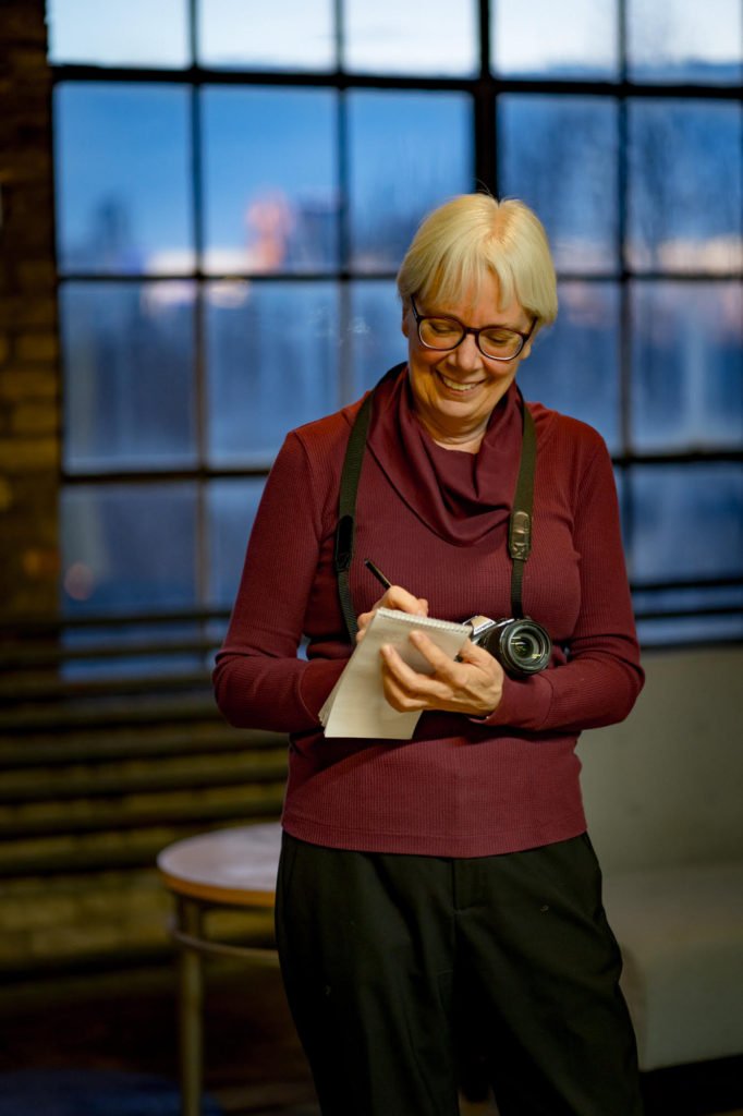 A smiling woman with blonde hair and glasses writes in a notepad, with a camera hanging around her neck.