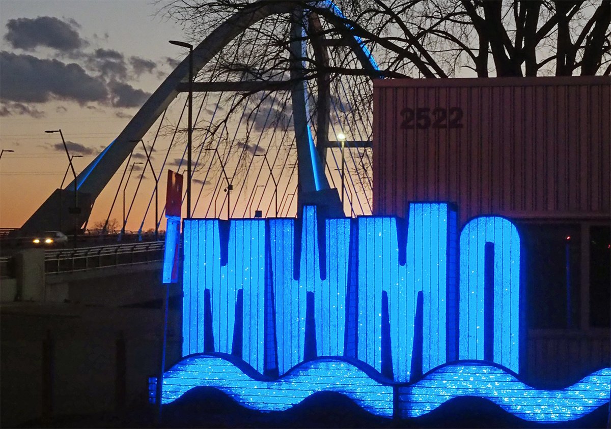 A blue neon lit sign saying MWMO in front of the Lowry Bridge at sunset.