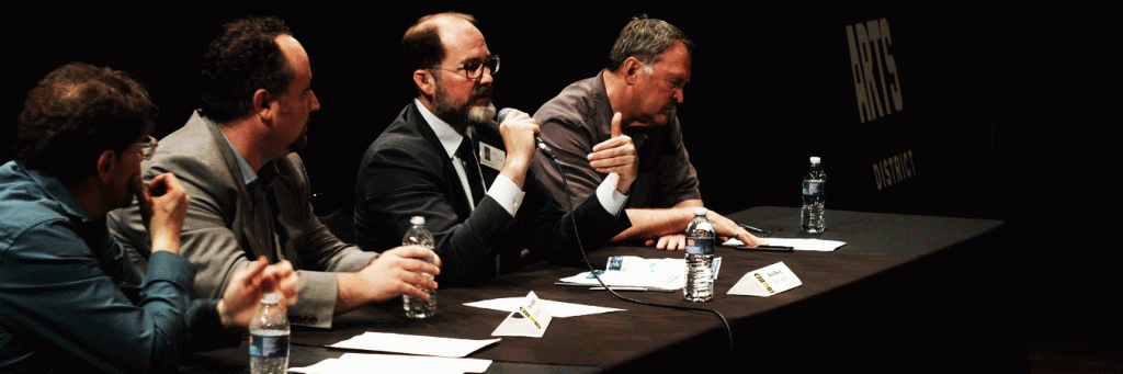 Four panelists sit at a table on a stage during an Arts District event, with one speaker holding a microphone and gesturing.