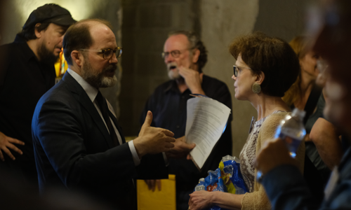 A man in a suit gestures while speaking to a woman holding a stack of papers and a water bottle in a crowded room.