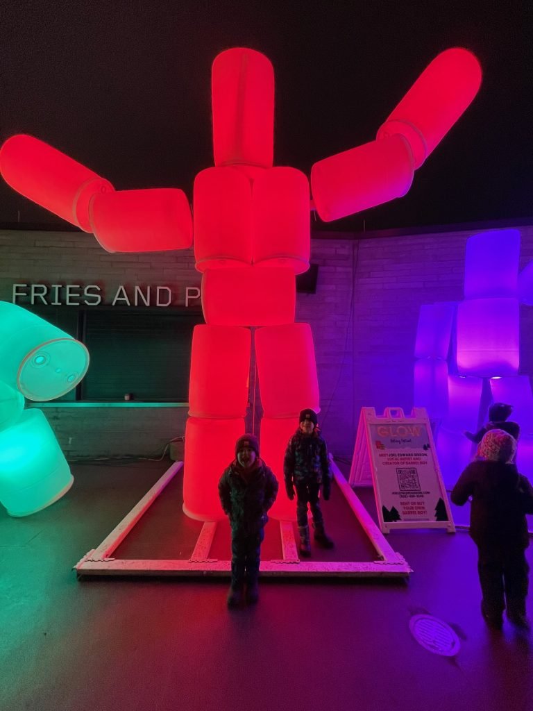 Two children stand in front of a large, glowing red inflatable figure made of connected cylinders, with colorful lit sculptures nearby.