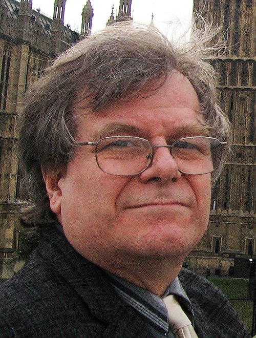 A portrait of a middle-aged man with glasses wearing a jacket and tie, in front of a stone building with gothic architecture.