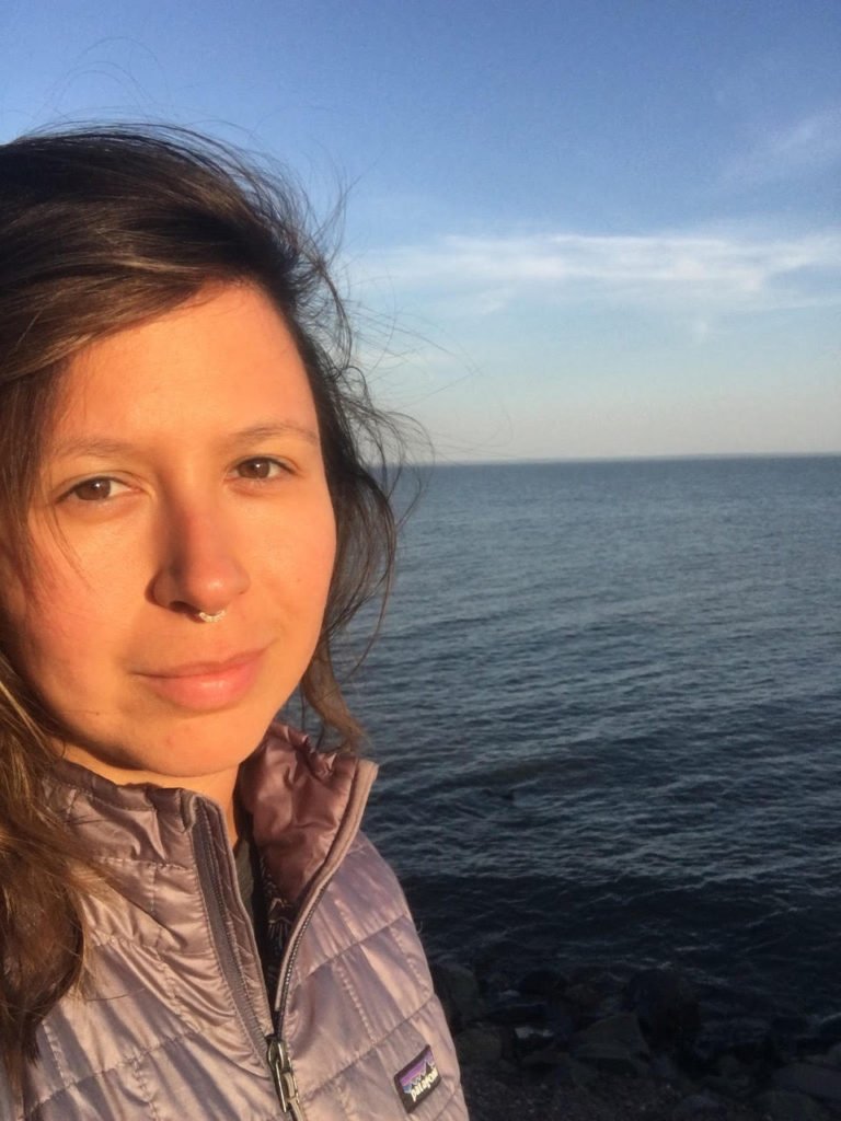 A woman in a puffer jacket stands by the ocean at sunset.