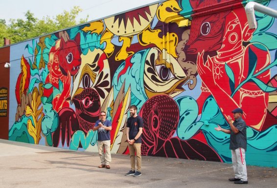Three people standing in front of a colorful mural of birds, cats and people.