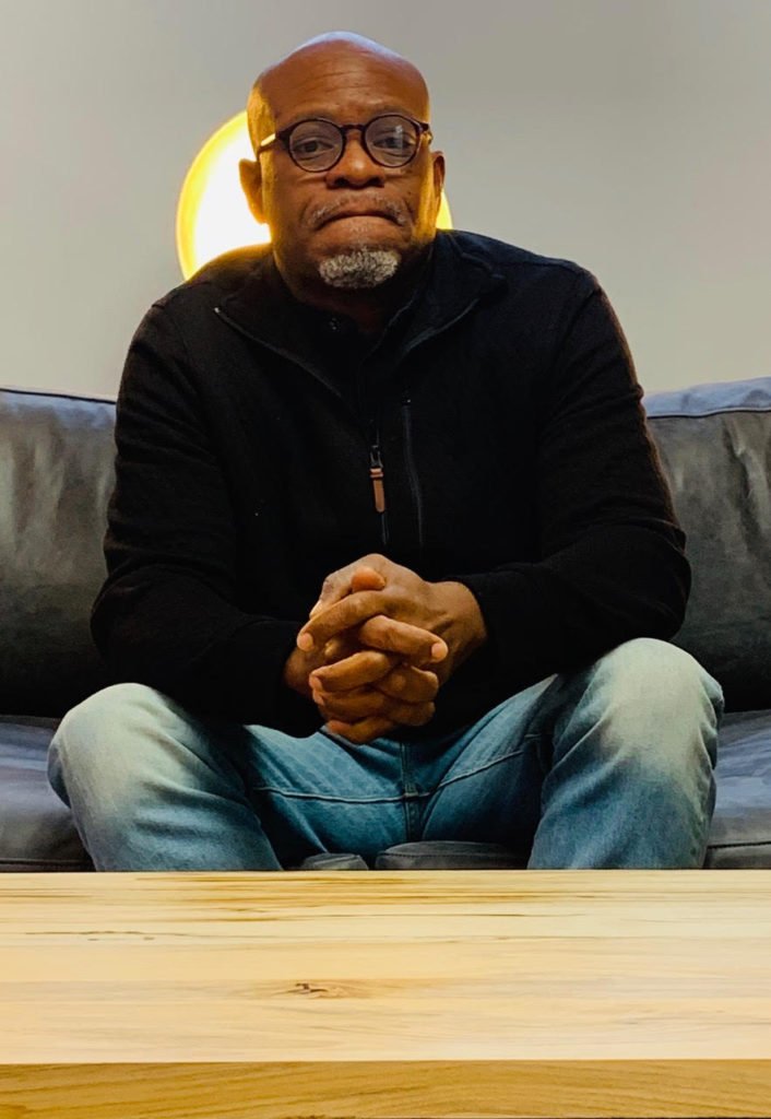 A Black man with a gray goatee and glasses, wearing a black zip-up sweater and jeans, sits with his hands clasped on a sofa.