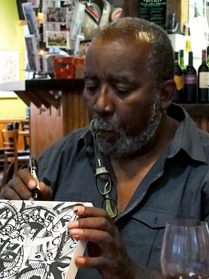 A man with a graying beard, wearing glasses around his neck, uses a black marker to draw on a small board with abstract white and black designs.