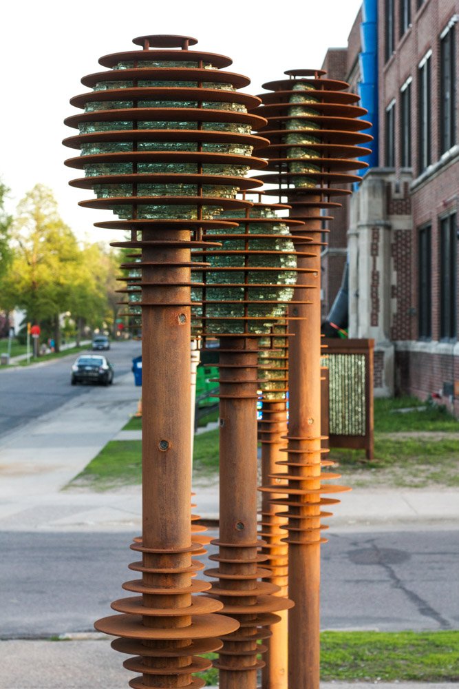 Three tall, rusted metal sculptures with stacked disc accents and textured glass spheres on top stand along a city sidewalk.