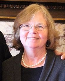 A smiling woman with shoulder-length blonde hair, wearing glasses, a pearl necklace, and a dark jacket.