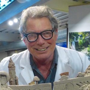 A smiling man in a white lab coat standing behind a clay model of a building.