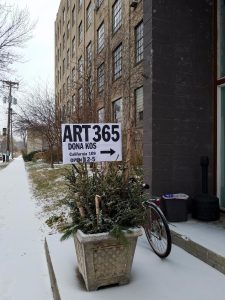 A sign on a sidewalk in the snow for ART365 by Dona Kos, indicating the location is at California 109 and open from 12-5.