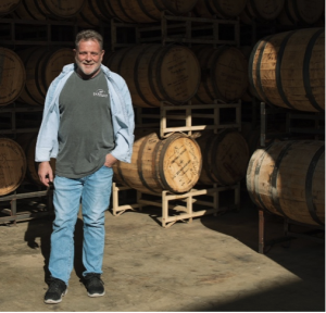 A smiling man in a gray t-shirt and jeans standing in a warehouse filled with stacked wooden barrels.